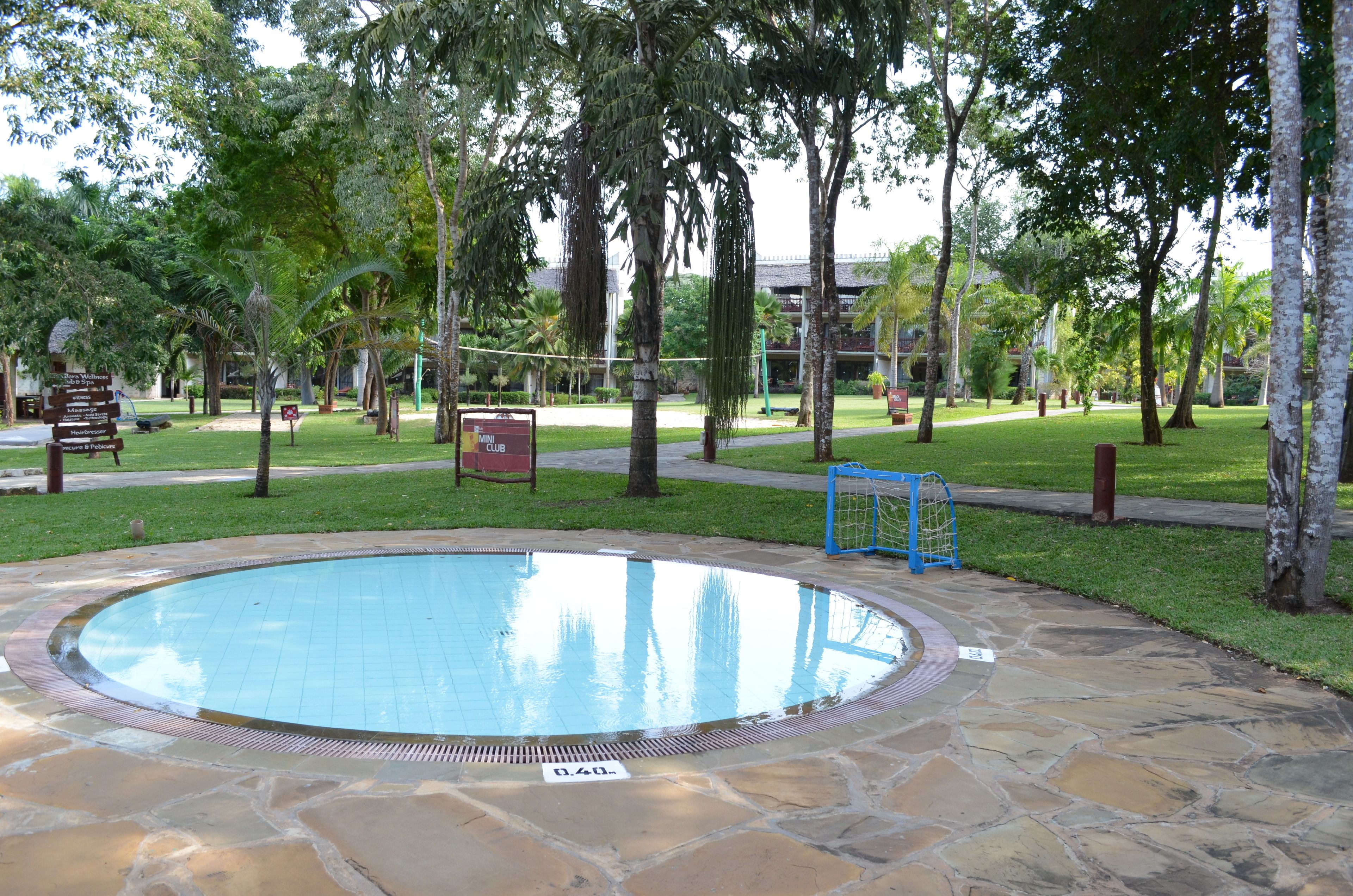 Uma piscina circular com borda de pedra, rodeada por um espaço gramado com árvores e uma cerca azul.