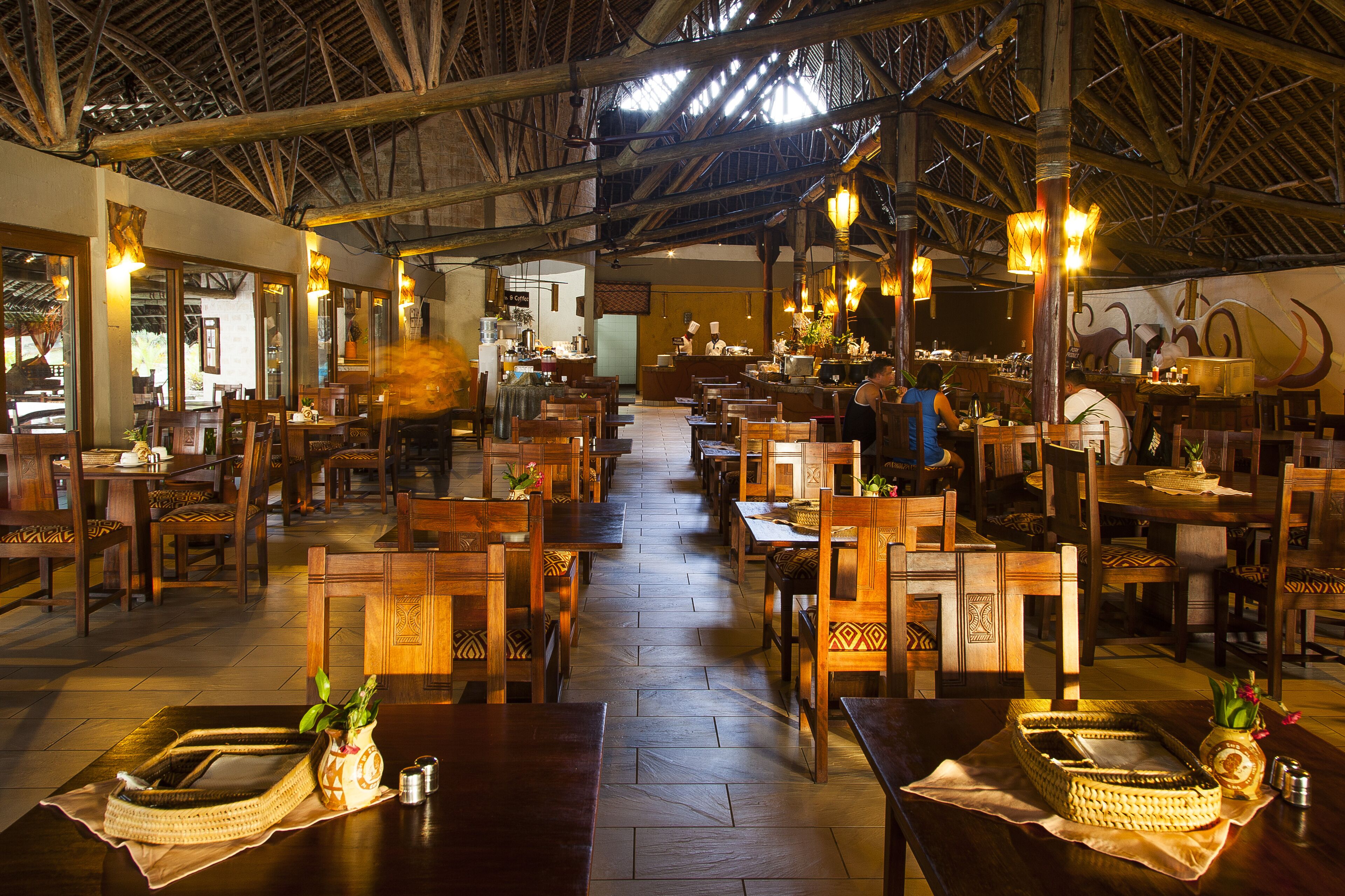 Un restaurante espacioso con mesas y sillas de madera, un techo alto con vigas a la vista, e iluminación cálida.