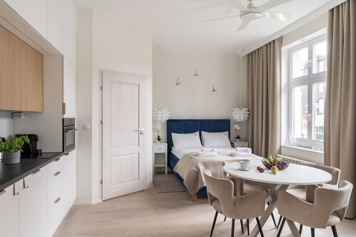 Quarto de hotel moderno com cozinha, cama com cabeceira azul, área de jantar com mesa e cadeiras, e uma janela grande com cortinas.