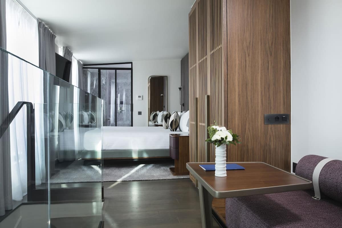 A modern hotel room with a glass partition, a bed, a wooden panel wall, a small table with a vase, and a sofa.