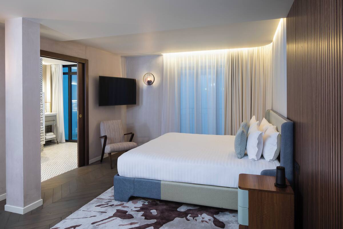 A modern hotel room with a large bed, a flat-screen TV mounted on the wall, a chair, and a wooden nightstand.