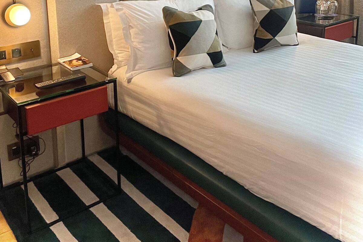 A hotel room with a bed, two bedside tables, a nightstand, a striped rug, and wall-mounted lights.
