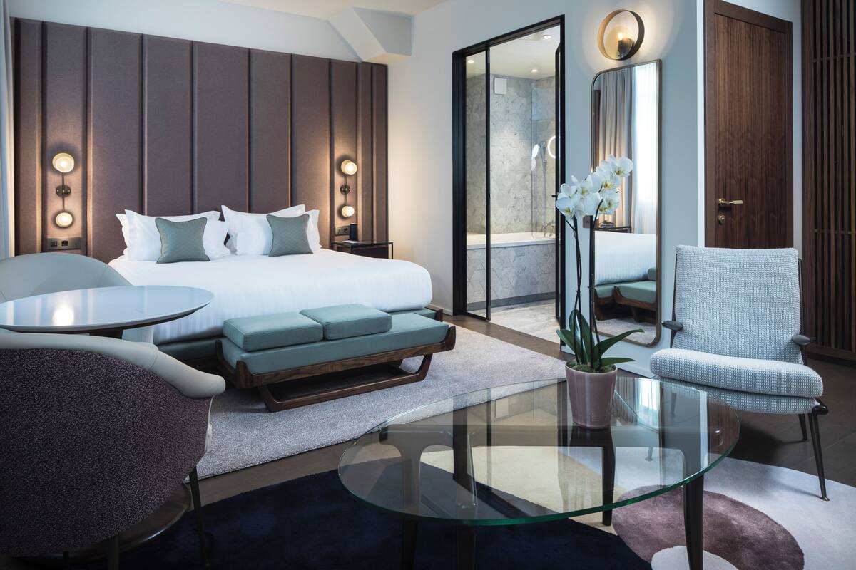 A modern hotel room with a large bed, a glass coffee table, a seating area with a chair and sofa, and a bathroom with a shower and mirror.