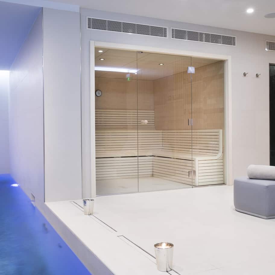 A modern spa room with a glass-enclosed sauna, a seating area with a towel, and a small pool.