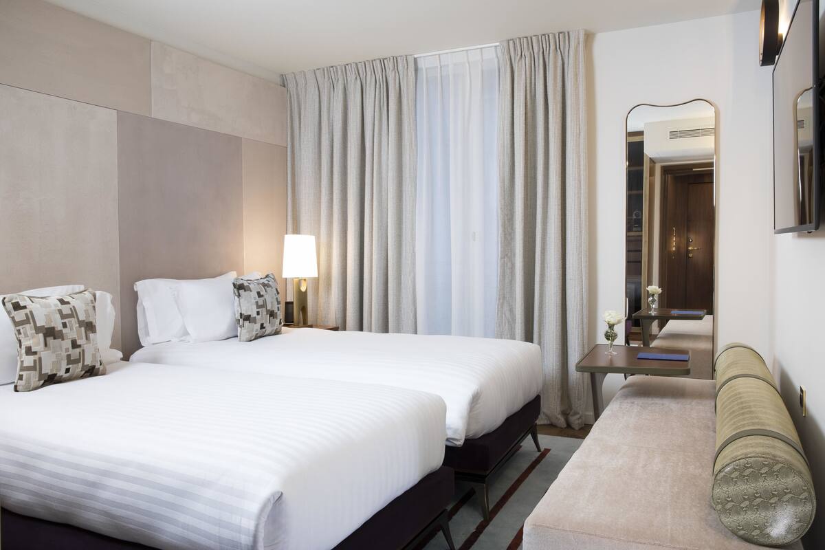 A hotel room with two beds, a bedside table with a lamp, a bench, a mirror, and a window with curtains.