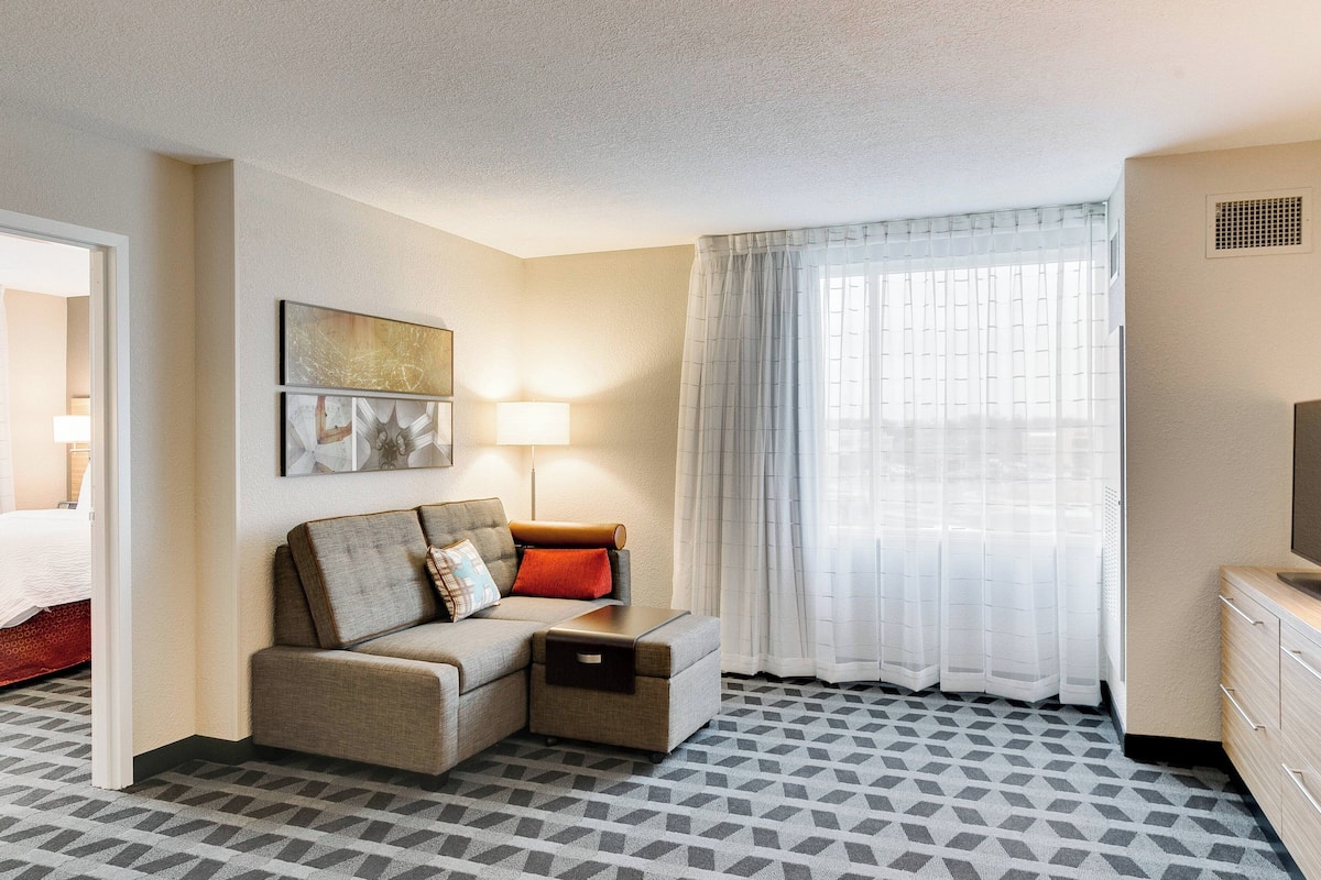Une chambre d’hôtel avec un canapé, une petite table, une lampe, une télévision et une fenêtre avec des rideaux.