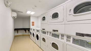 A laundry room with multiple rows of white washing machines and dryers, a ventilation system, and a sign indicating the room's purpose.