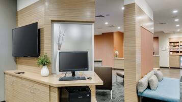 A reception desk with a flat-screen TV, computer, keyboard, and printer. There is a plant on the desk, a sofa with cushions, and a patterned carpet.