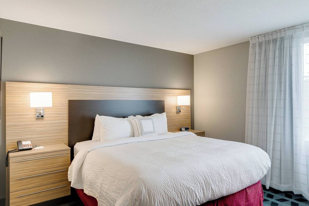 Une chambre d’hôtel avec un grand lit, deux tables de chevet, une petite table, une lampe et une fenêtre avec des rideaux.