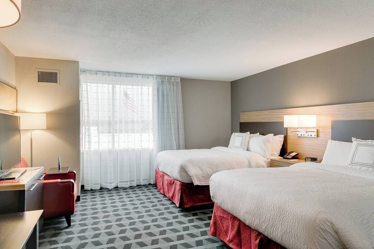 Une chambre d’hôtel avec deux lits, un bureau, une chaise, une lampe et une fenêtre avec des rideaux.