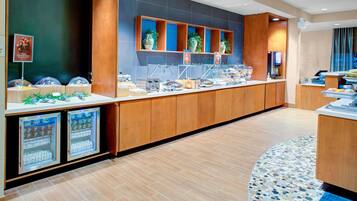 A modern hotel breakfast area with a long counter, refrigerated display cases, and potted plants.