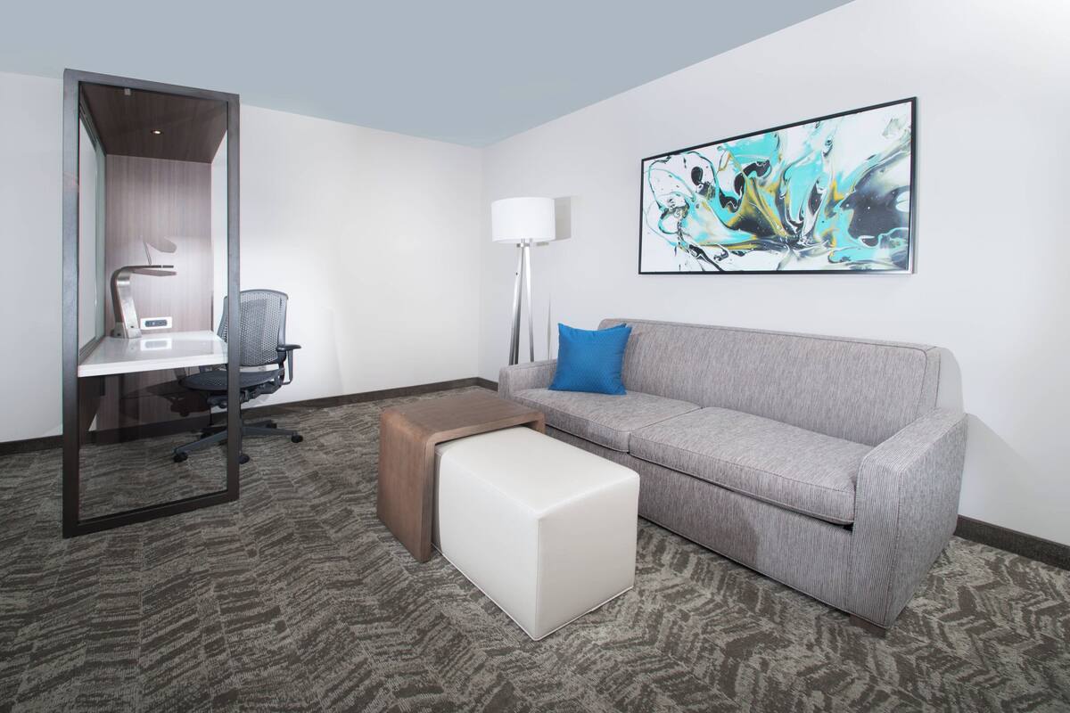 A modern hotel room with a grey sofa, a small white ottoman, a desk with a chair, a standing lamp, and a large abstract painting on the wall.