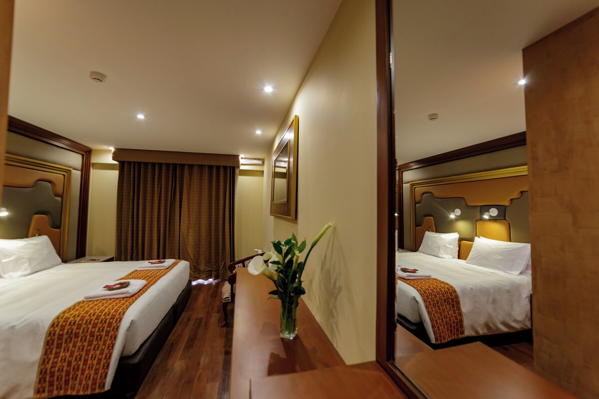 A hotel room with two beds, a wooden floor, a mirror, and a vase with flowers.