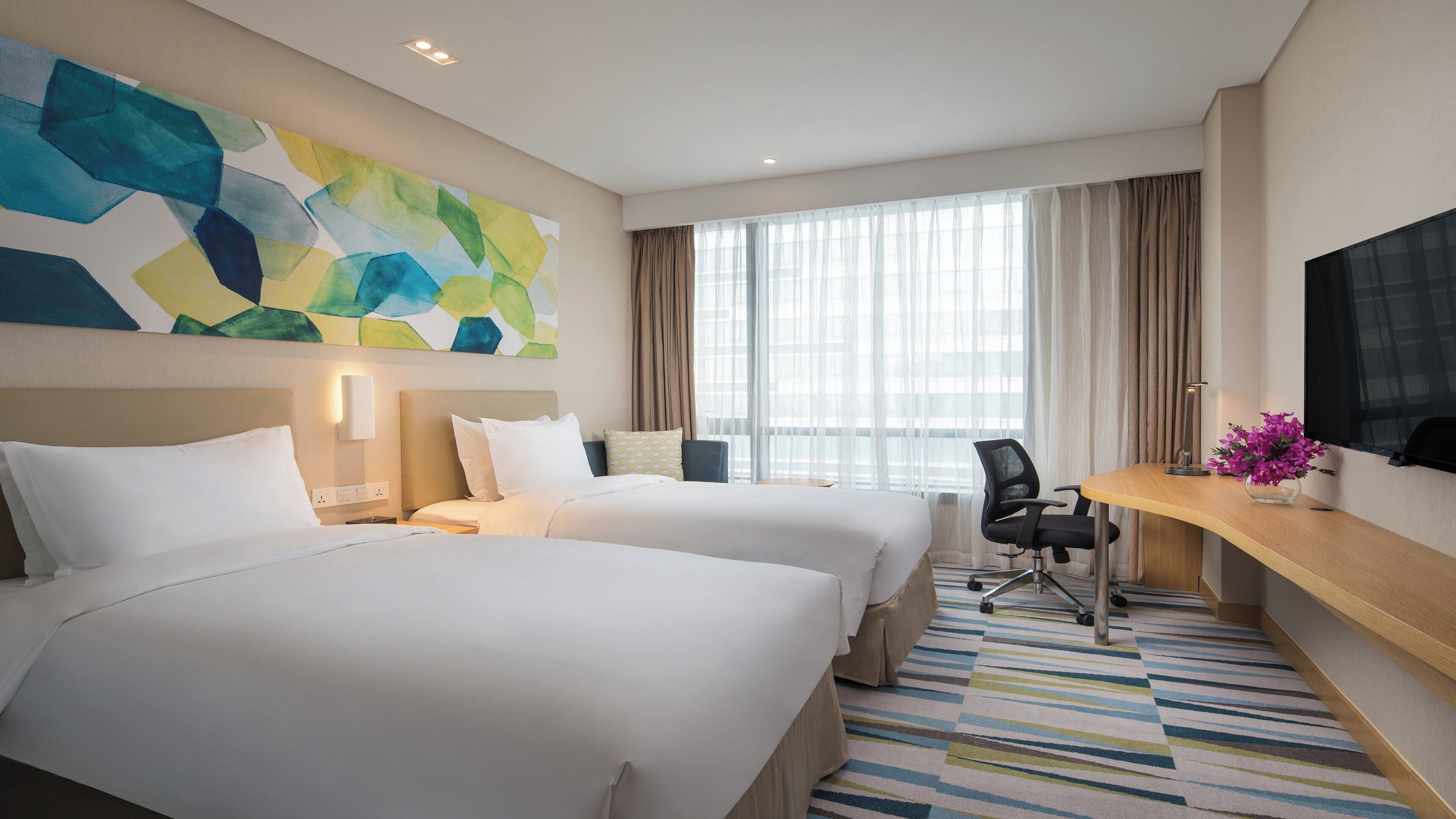 A hotel room with two beds, a desk, a chair, a TV, and a large window with curtains.