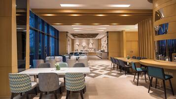 A modern hotel dining area with a long table, multiple chairs, and a kitchen in the background.