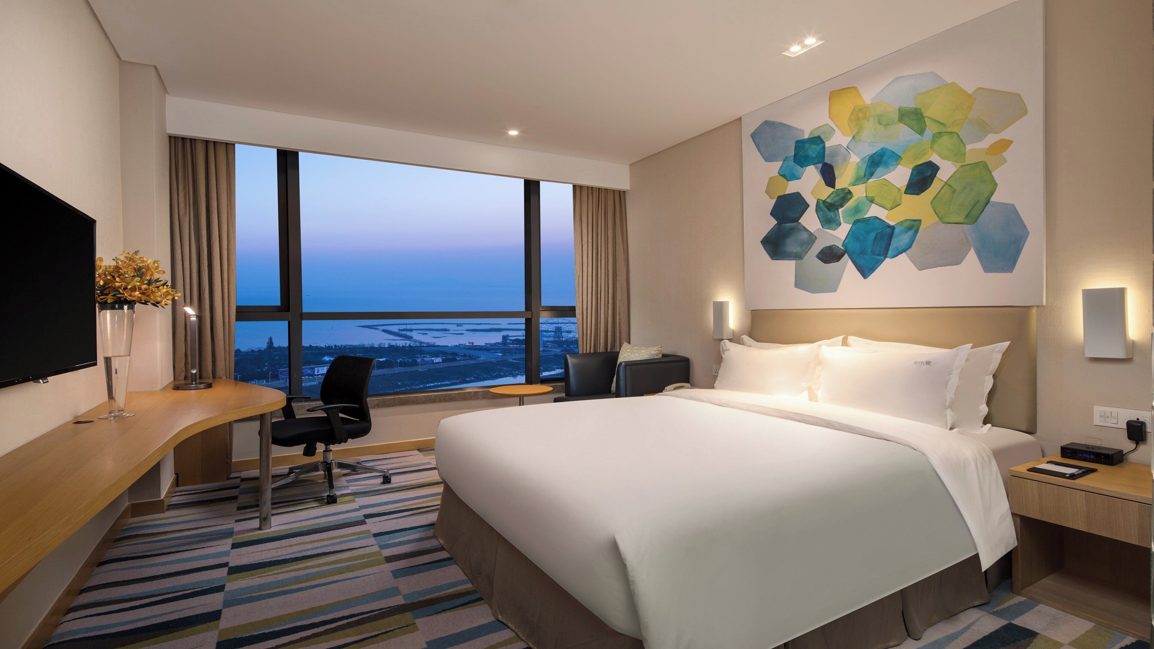 Habitación de hotel con una cama grande, un escritorio, una silla, un televisor y vistas a la ciudad a través de la ventana.