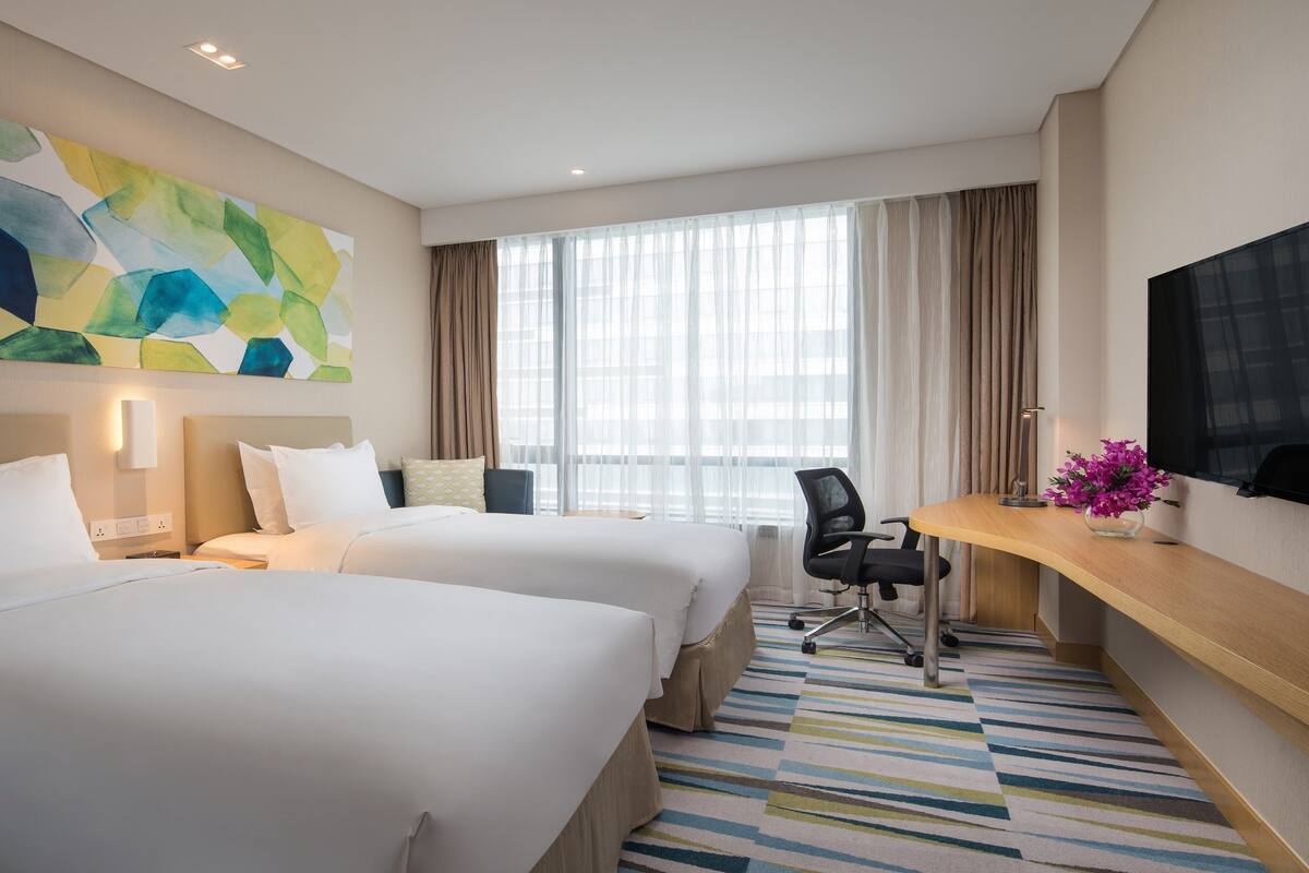 A hotel room with two beds, a desk, a chair, a TV, and a large abstract painting on the wall.
