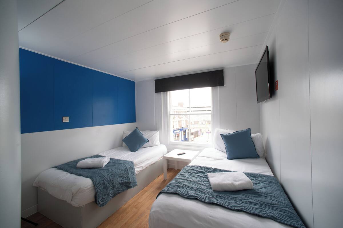 A modern hotel room with two beds, a small table, a window with a view, and a mounted TV.