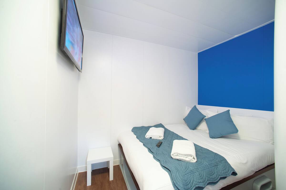 A neatly made bed with a blue headboard, white bedding, and two pillows. A folded towel is placed on the bed, and a remote control is visible on the bed's surface.