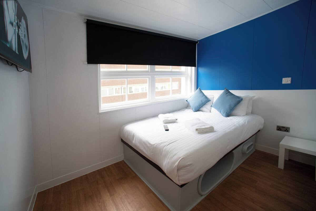 A bedroom with a bed, a window with blinds, a white bench, and a wall with a blue panel.