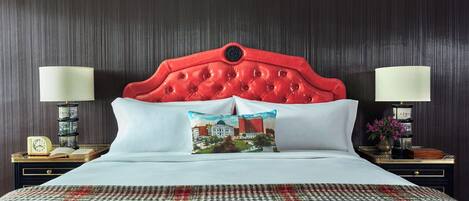 Habitación de hotel con cabecera tapizada de rojo, ropa de cama blanca, una manta de cuadros y dos mesitas de noche con lámparas y relojes.