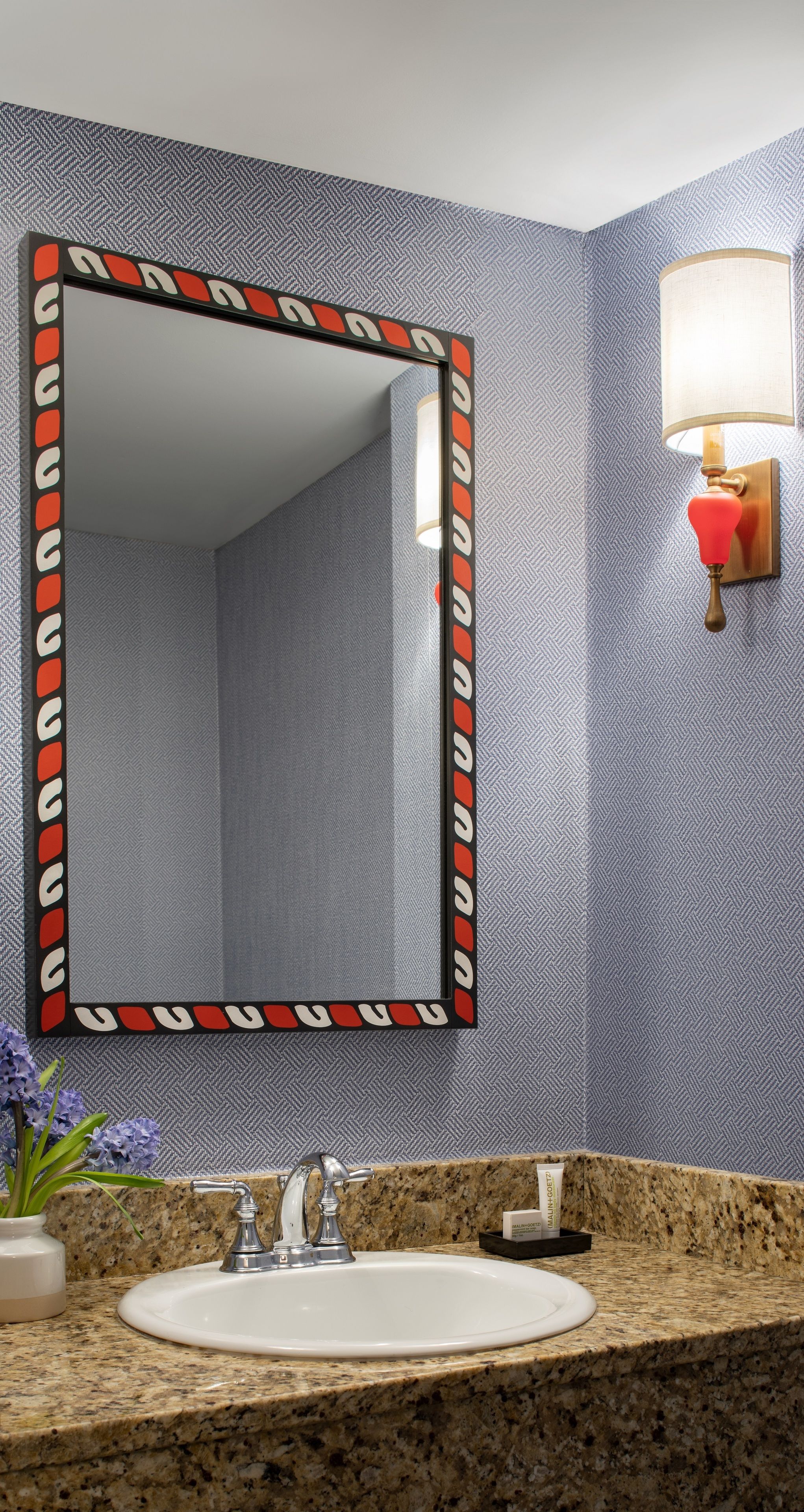 A bathroom with a granite countertop, a white sink, a mirror with a decorative frame, and a wall-mounted light fixture.