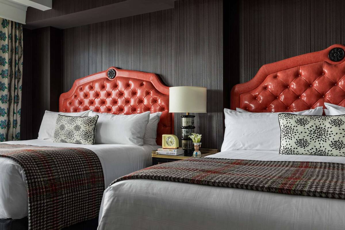 Ein Hotelzimmer mit zwei Betten, jeweils mit rotem Kopfteil, weißer Bettwäsche und einer gemusterten Decke. Ein Nachttisch mit Lampe und kleiner Uhr ist zu sehen.