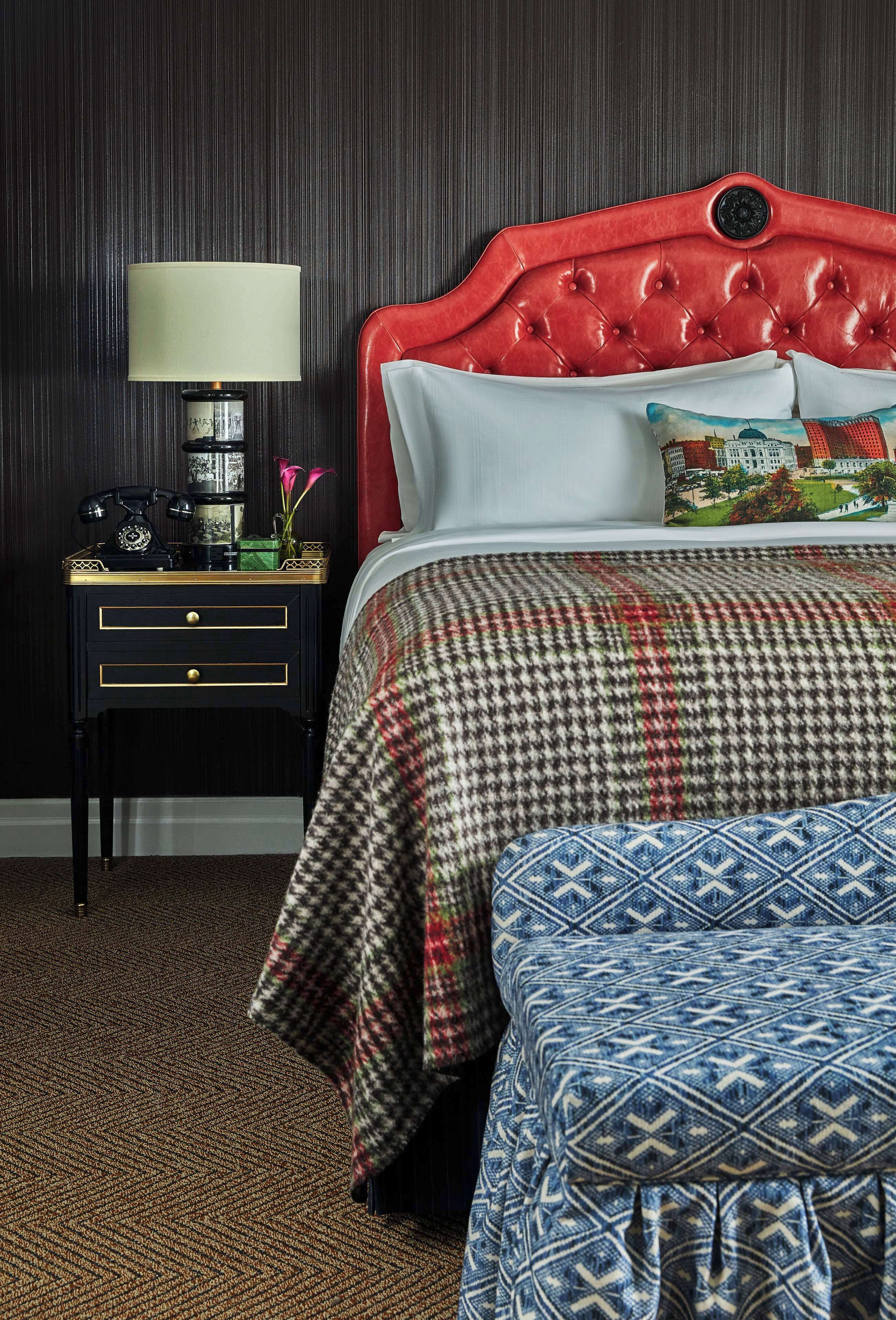 Habitación de hotel con cabecera roja, colcha estampada, reposapiés azul con diseño, mesita de noche con lámpara y un jarrón decorativo.