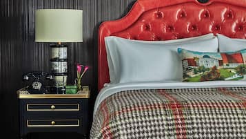 Habitación de hotel con cabecera roja, colcha estampada, reposapiés azul con diseño, mesita de noche con lámpara y un jarrón decorativo.