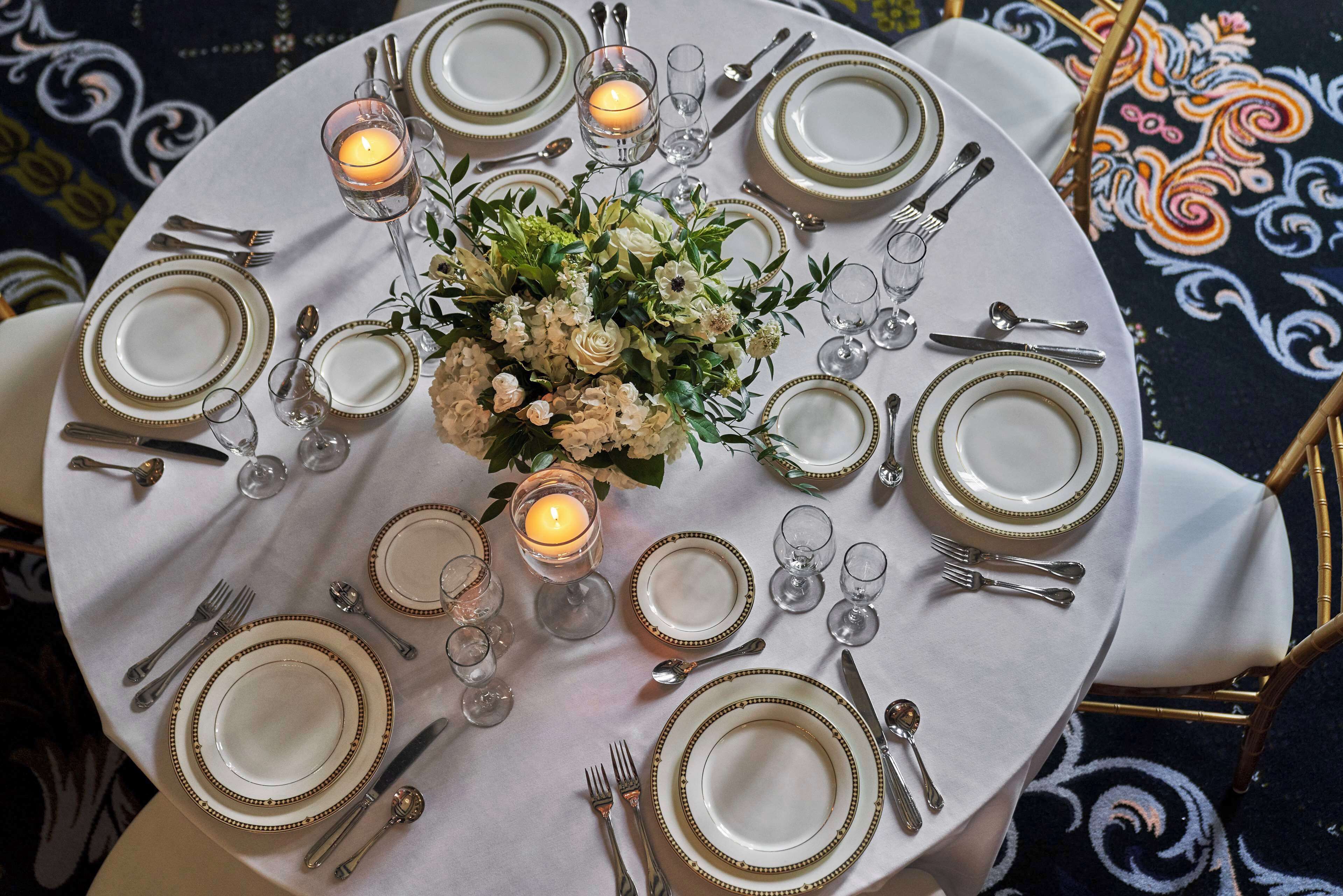 Une table ronde, dressée pour le dîner, est ornée d’assiettes blanches, de couverts à bord doré et de verres transparents. Un arrangement floral et des bougies en sont le point central.