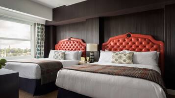 A hotel room with two beds, a window with curtains, a bedside table with a lamp, and a view of a cityscape.