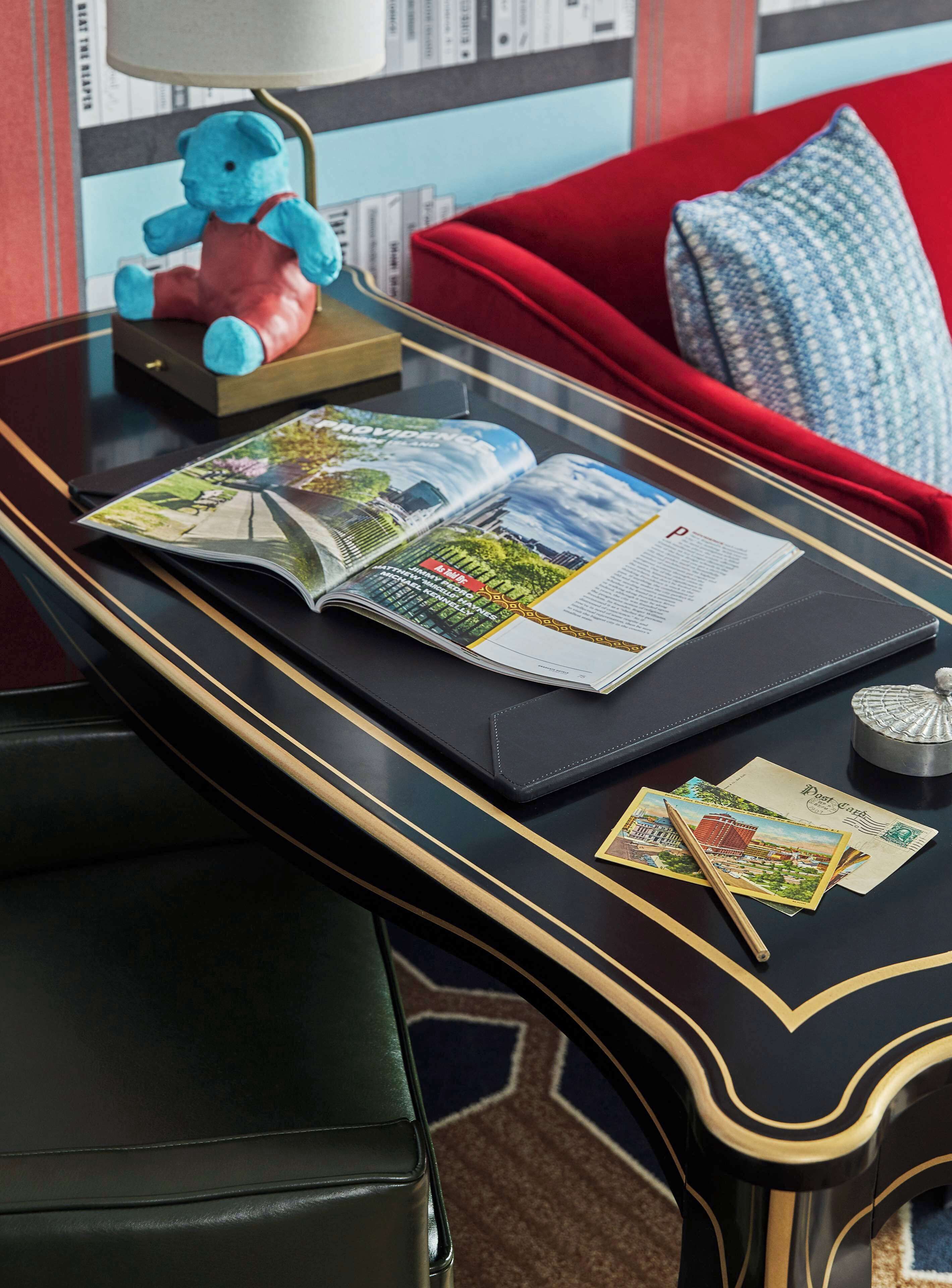 Una mesa negra con detalles dorados, una revista, postales, un bolígrafo y una lámpara pequeña.