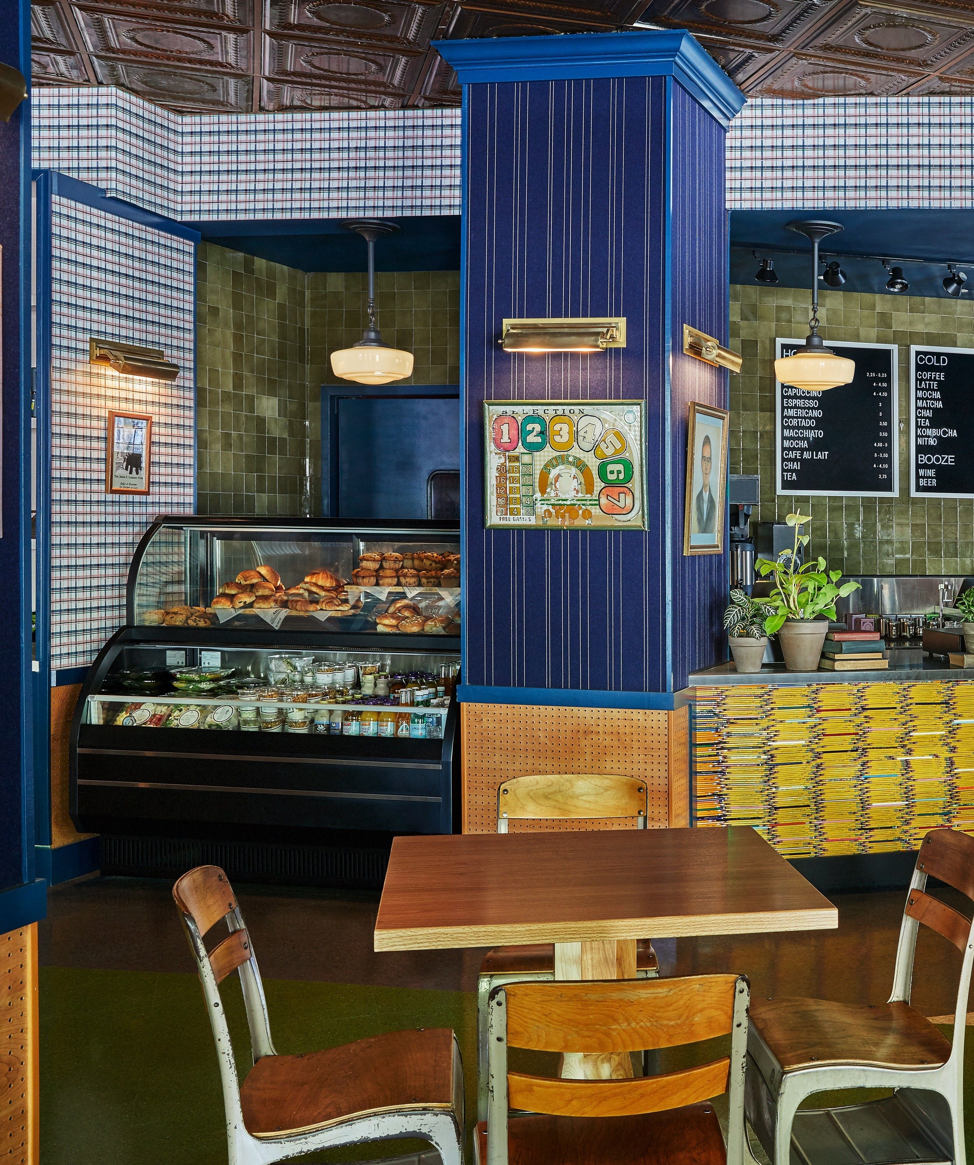 Ein Café mit einer blau-grün karierten Wand, einer Auswahltafel, einer Speisekarte und einer Vitrine mit Gebäck.