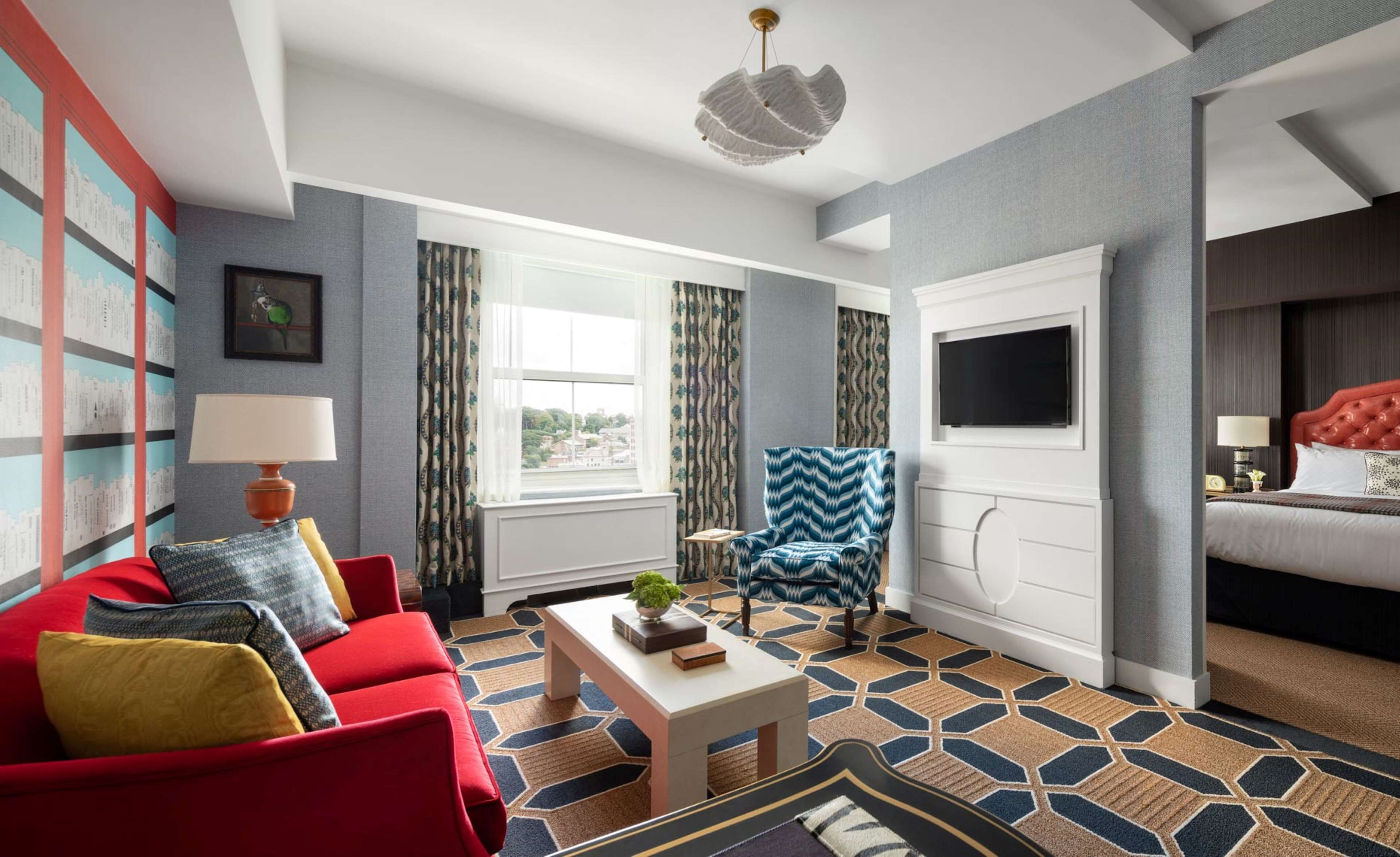 Une chambre d’hôtel moderne avec un canapé rouge, un fauteuil à motifs, une table de chevet blanche, une grande fenêtre et un téléviseur à écran plat fixé au-dessus du lit.