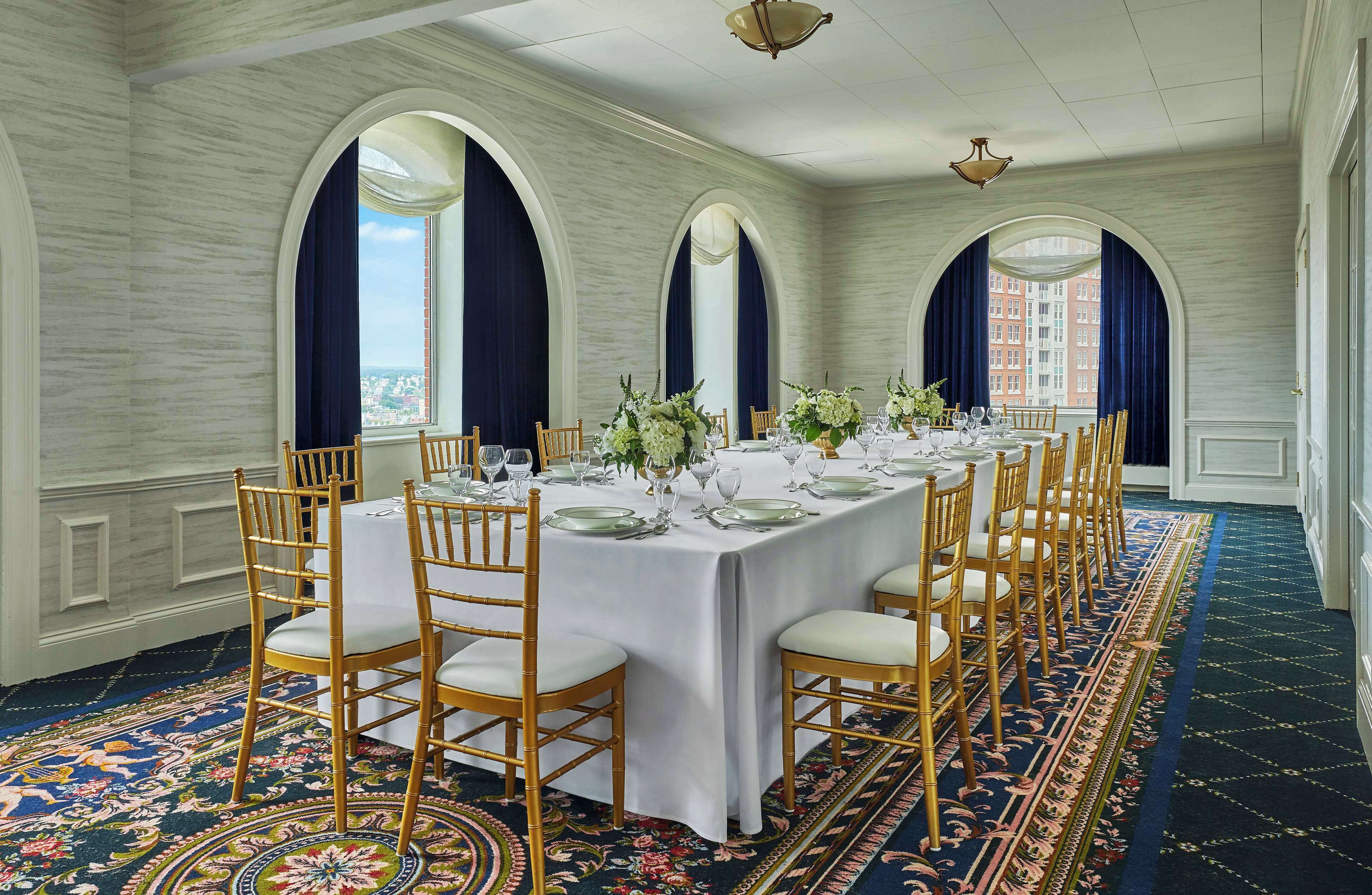A long dining table set for a formal event with chairs, a patterned carpet, and large windows with curtains.