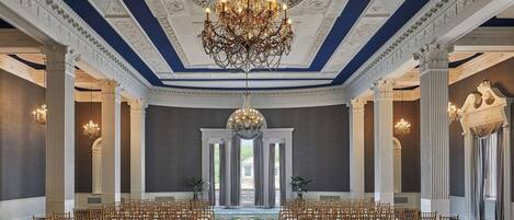 A grand hall with ornate ceiling and chandeliers, rows of wooden chairs, and a patterned carpet.