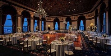 A grand ballroom with multiple round tables, elegant chandeliers, and large windows offering city views.