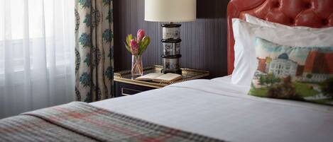 Une chambre d’hôtel avec un lit, une lampe de chevet, un vase de fleurs et un oreiller orné d’un motif représentant un paysage urbain.