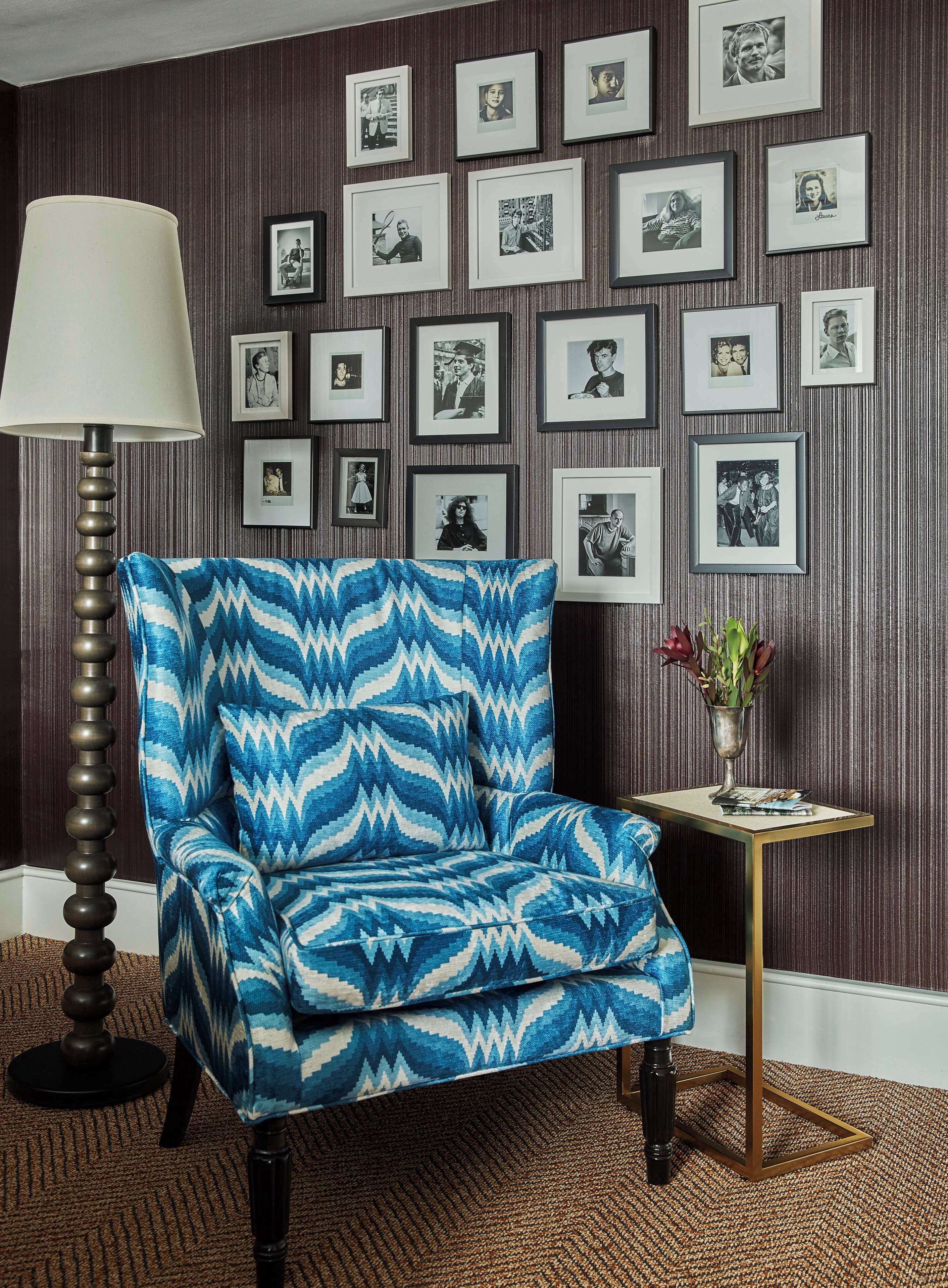 A room with a patterned armchair, a floor lamp, and a wall adorned with framed photographs.