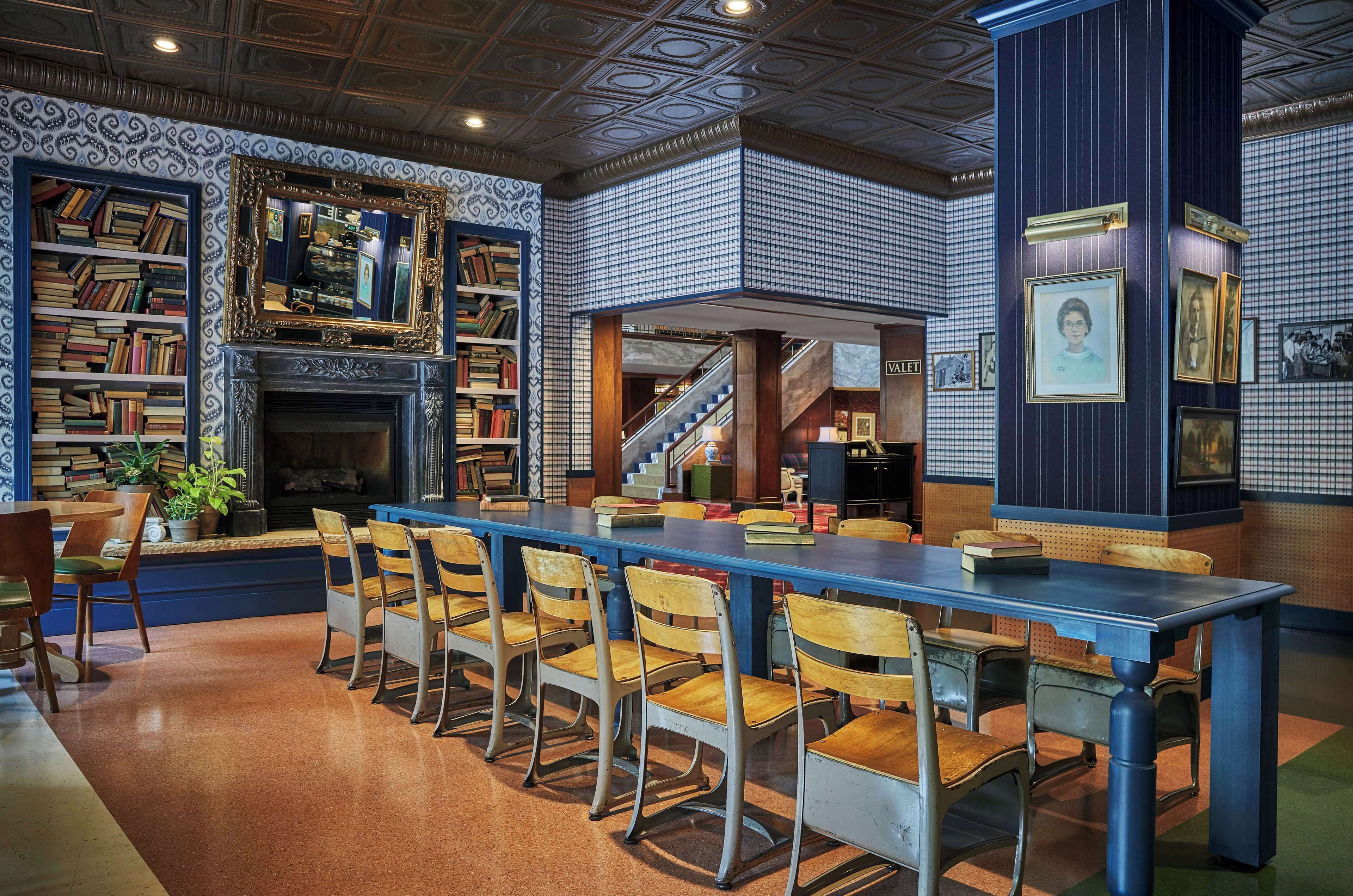 A library with bookshelves, a fireplace, a long table with chairs, and a staircase.