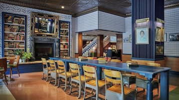 A library with bookshelves, a fireplace, a long table with chairs, and a staircase.