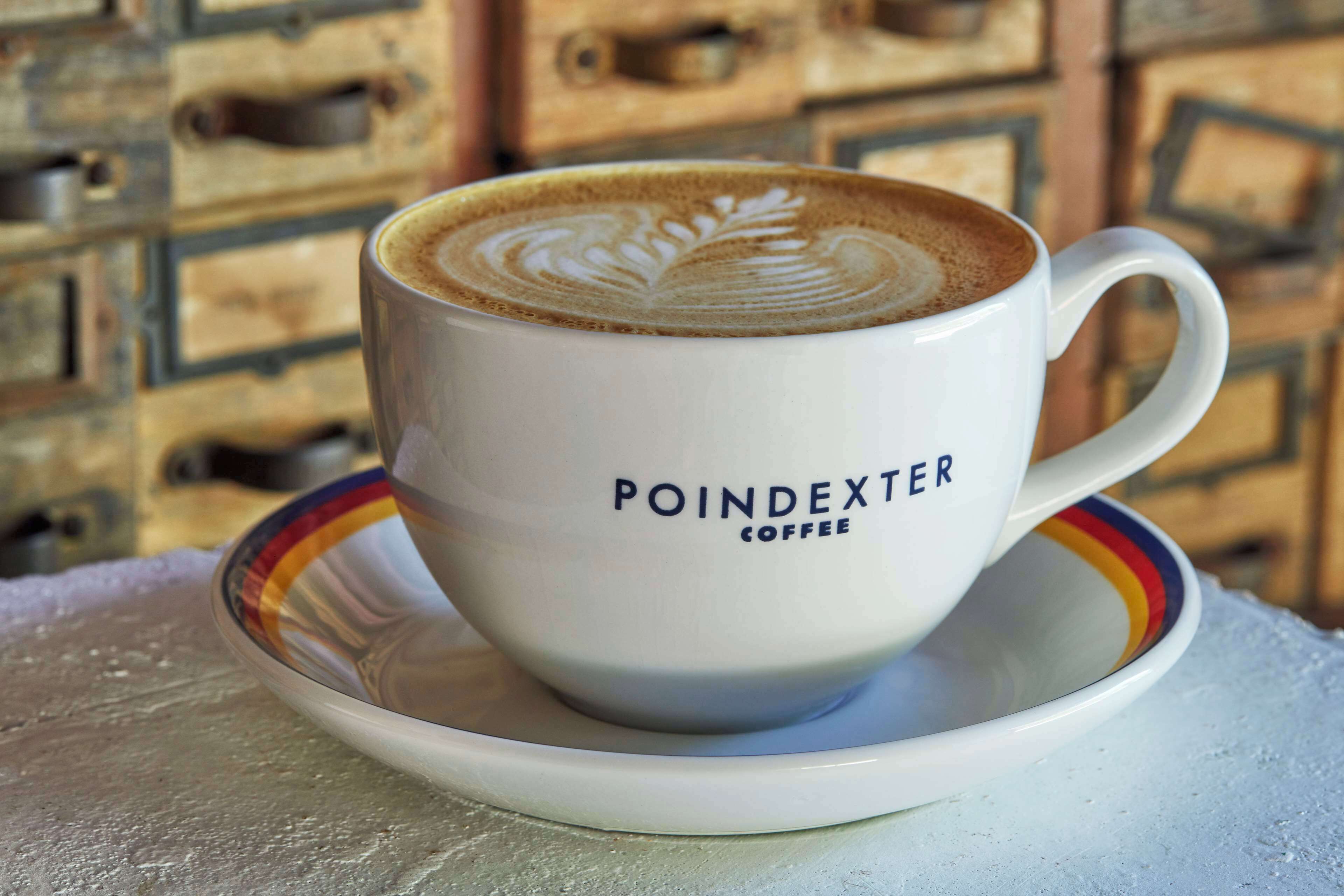 A white coffee cup with a leaf design on the foam, labeled "POINDEXTER COFFEE," on a saucer.