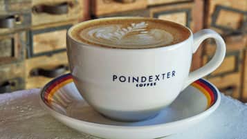 A white coffee cup with a leaf design on the foam, labeled "POINDEXTER COFFEE," on a saucer.