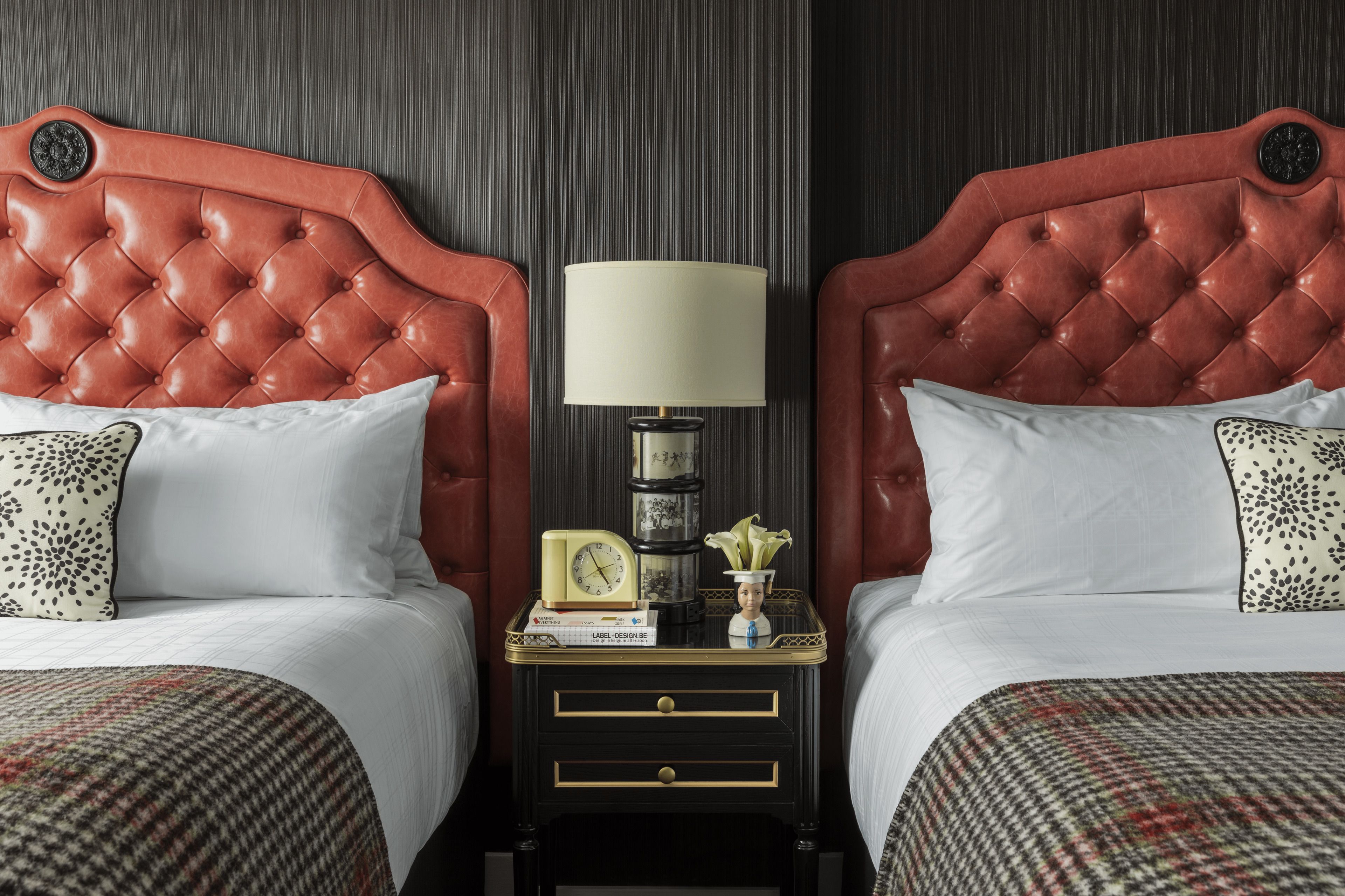 A hotel room with two beds, each with a red headboard, white bedding, and patterned pillows. There is a nightstand between the beds with a lamp, a clock, and a small vase with flowers.