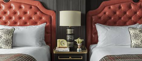 A hotel room with two beds, each with a red headboard, white bedding, and patterned pillows. There is a nightstand between the beds with a lamp, a clock, and a small vase with flowers.