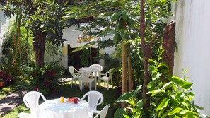 Une terrasse extérieure avec une table ronde et des chaises blanches, entourée d’une végétation luxuriante et d’un mur blanc.