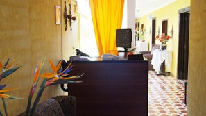 Un comptoir d’accueil avec un téléphone, un écran d’ordinateur et une plaque nominative. On aperçoit, en arrière-plan, des plantes décoratives et un rideau jaune.