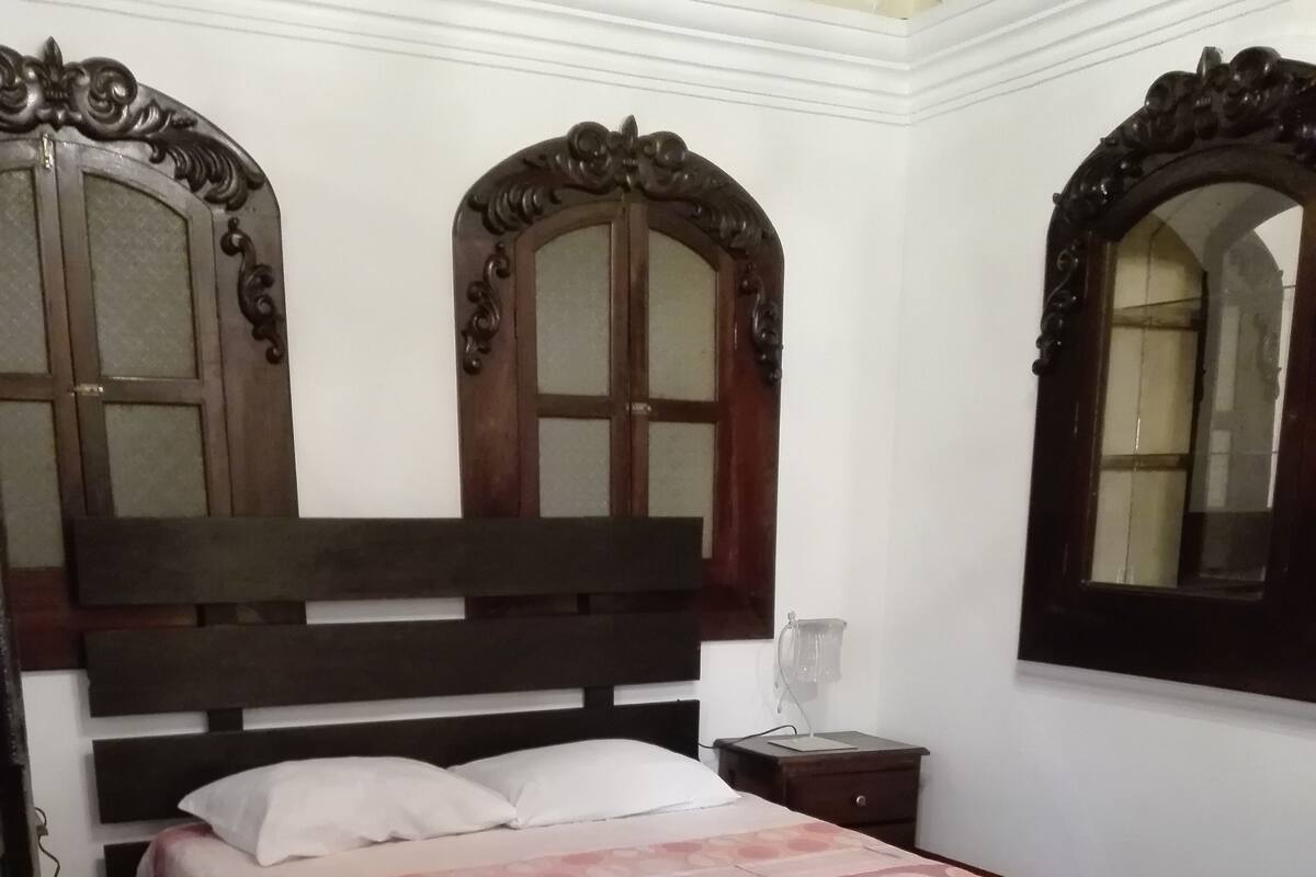 A bedroom with a bed, wooden headboard, two windows with decorative frames, a bedside table with a lamp, and a patterned floor.