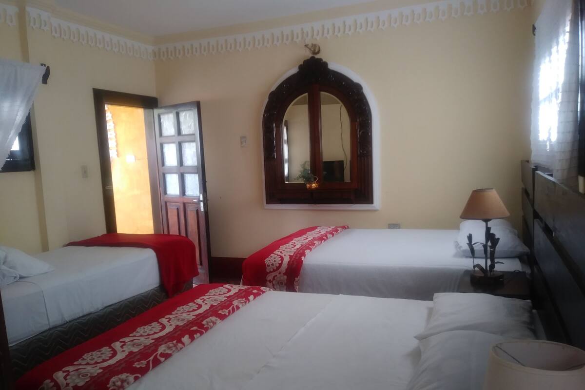 A hotel room with two single beds, a large mirror, a bedside lamp, and a window with curtains.