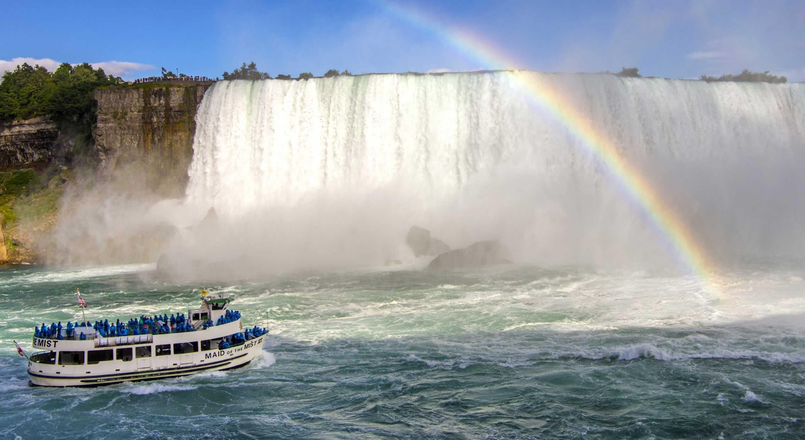 A boat named 'MAID OF THE MIST' with passengers, a large waterfall, and a rainbow.